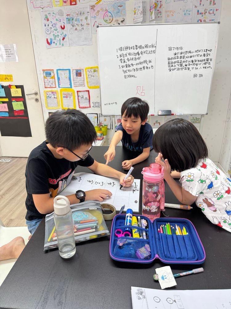 Children working together on a Chinese writing activity in a classroom, discussing characters around a shared worksheet with colourful stationery on the table.